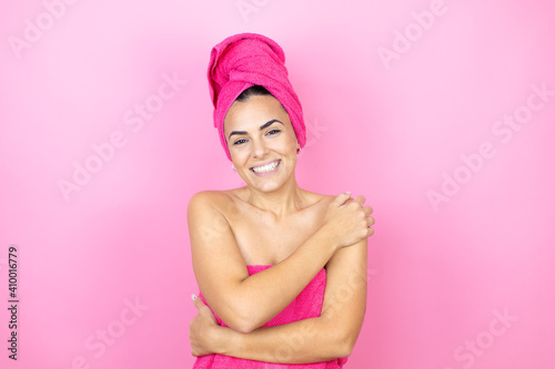Young beautiful woman wearing shower towel after bath standing over isolated pink background hugging oneself happy and positive, smiling confident
