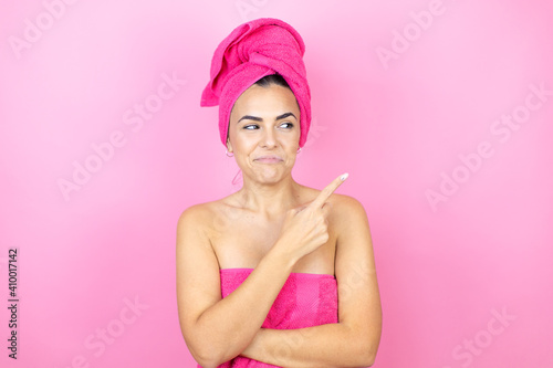 Young beautiful woman wearing shower towel after bath standing over isolated pink background confused and pointing with hand and finger to the side