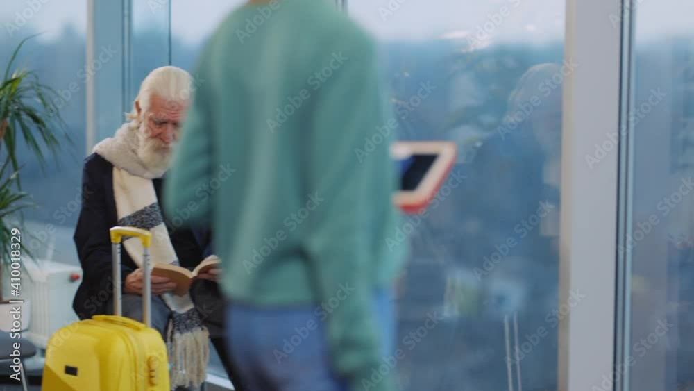 Old caucasian man reading book in passenger waiting room. Young afro-american attractive woman with tablet and suitcase entering departure lounge. Airport. Old caucasian man reading book in passenger waiting room. Young afro-american attractive woman with tablet and suitcase entering departure lounge. Airport.
