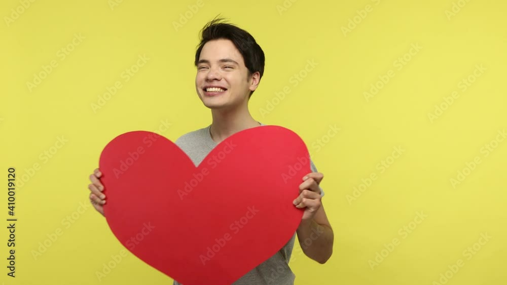Extremely happy young adult man holding big red paper heart and smiling, preparing gift to saint valentines day, happy to receive greeting card. Indoor studio shot isolated on yellow background Extremely happy young adult man holding big red paper heart and smiling, preparing gift to saint valentines day, happy to receive greeting card. Indoor studio shot isolated on yellow background