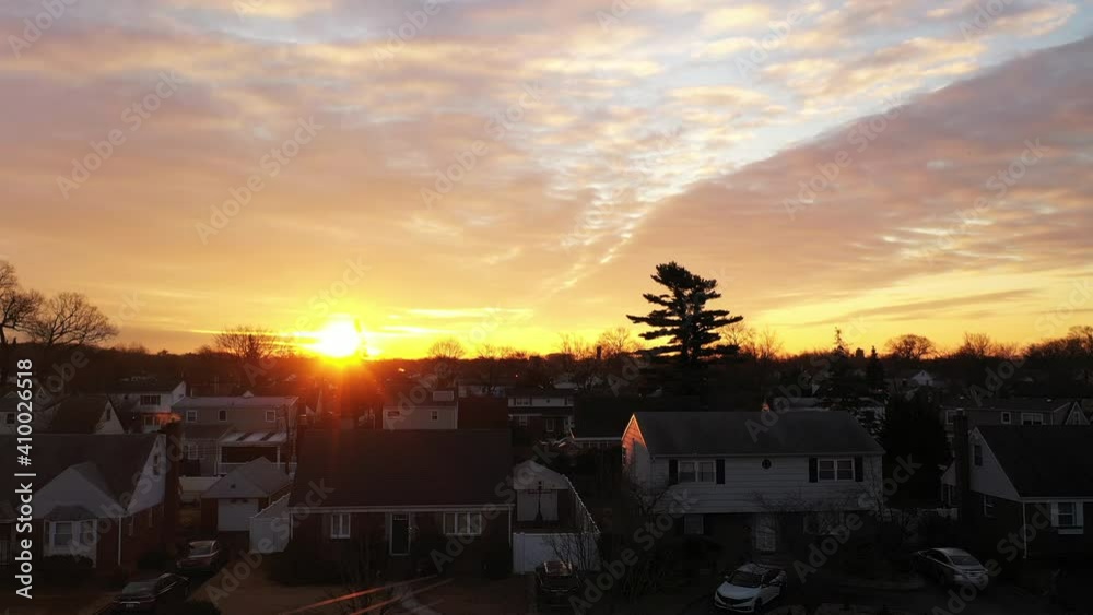 An drone view of a Long Island neighborhood during a golden sunrise with clouds and blue skies. The camera boom up pointing towards the sun on the horizon.