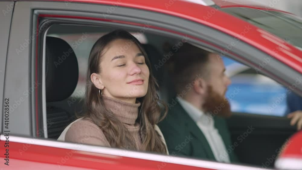 Young brunette Caucasian woman with brown eyes closing car door and talking to blurred man sitting on driver's seat. Portrait of positive couple choosing new vehicle in auto dealership. Wealth concept