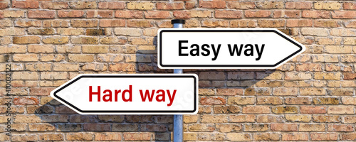 signpost with two arrow-shaped signs with the words HARD WAY and EASY WAY in opposite direction in front of a brick wall