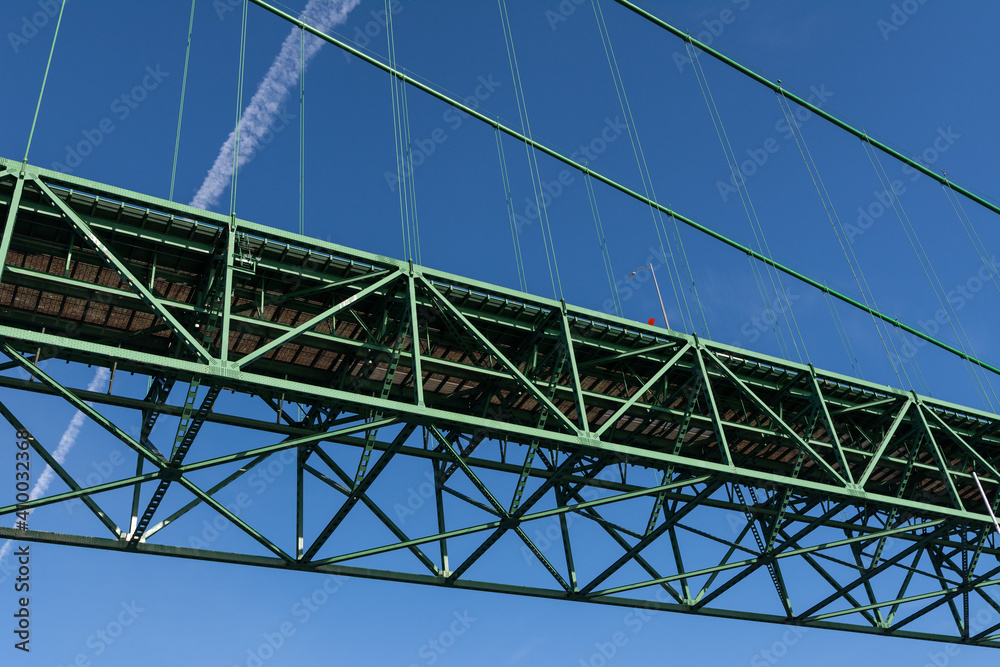 Closeup of a section of green metal suspension bridge against a bright blue sky; contrail from plane