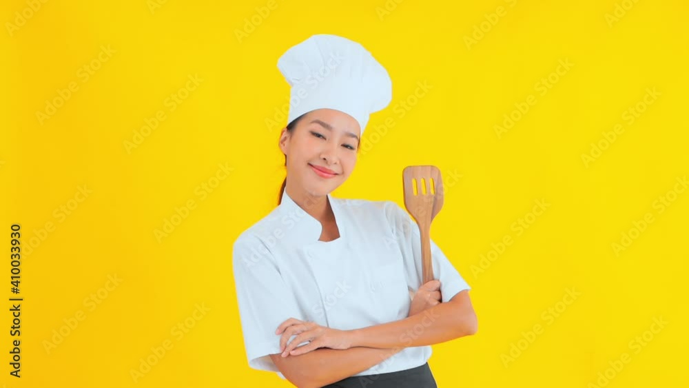 An attractive female chef in a traditional chef's uniform is standing against a solid background, arms folded, wooden spatula in hand. An attractive female chef in a traditional chef's uniform is standing against a solid background, arms folded, wooden spatula in hand.