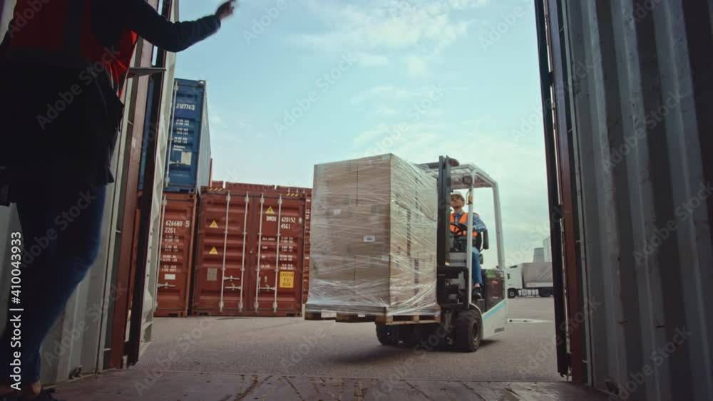 Forklift Driver Loading a Shipping Cargo Container with a Full Pallet with Carboard Boxes in Logistics Operations Port Terminal. Female Industrial Supervisor and Safety Inspector Helping the Process.