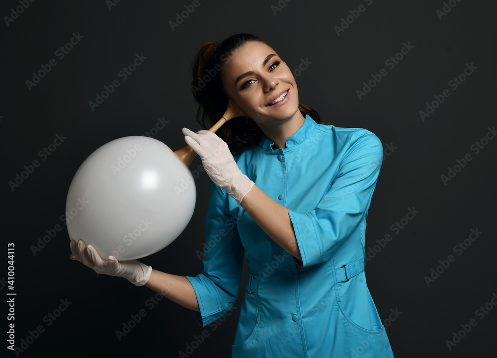 Smiling pretty brunette woman doctor gynecologist in latex gloves and medical uniform gown uses stethoscope tube listening air balloon over dark background. Gynecology, healthcare, medicare concept Smiling pretty brunette woman doctor gynecologist in latex gloves and medical uniform gown uses stethoscope tube listening air balloon over dark background. Gynecology, healthcare, medicare concept