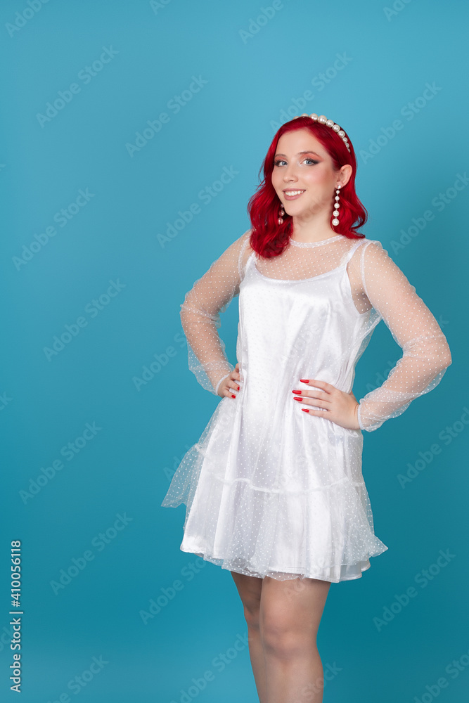 joyful contented beautiful young woman with red hair and hands on waist in white silk mesh dress isolated on blue background. joyful contented beautiful young woman with red hair and hands on waist in white silk mesh dress isolated on blue background.