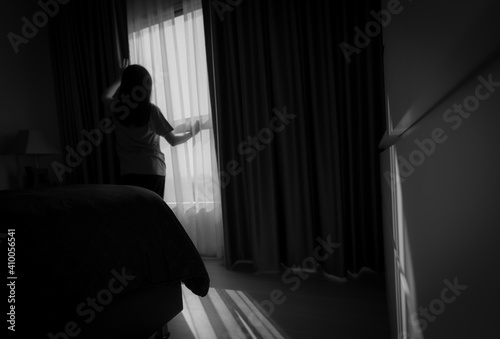 Rear view of woman holding curtain and looking out of glass window. Depressed woman from lock down and quarantine from coronavirus pandemic. Mental health and dementia. Thoughtful woman in bedroom.