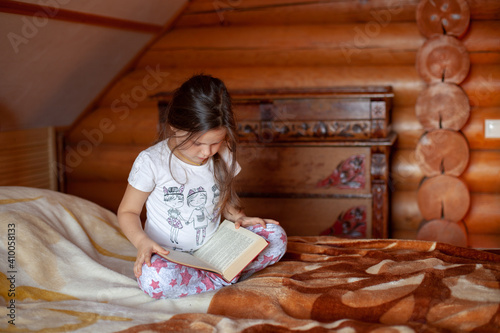 a passionate five-year-old girl sits cross-legged on a bed and reads a book in the bedroom of a log cabin in the village.