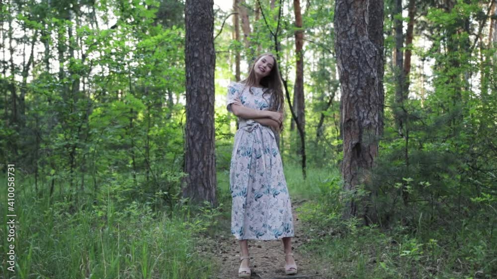 A beautiful woman smiles as she walks along a path surrounded by foliage and trees. Front view of a woman in a dress walking in a summer Park. Slow motion A beautiful woman smiles as she walks along a path surrounded by foliage and trees. Front view of a woman in a dress walking in a summer Park. Slow motion