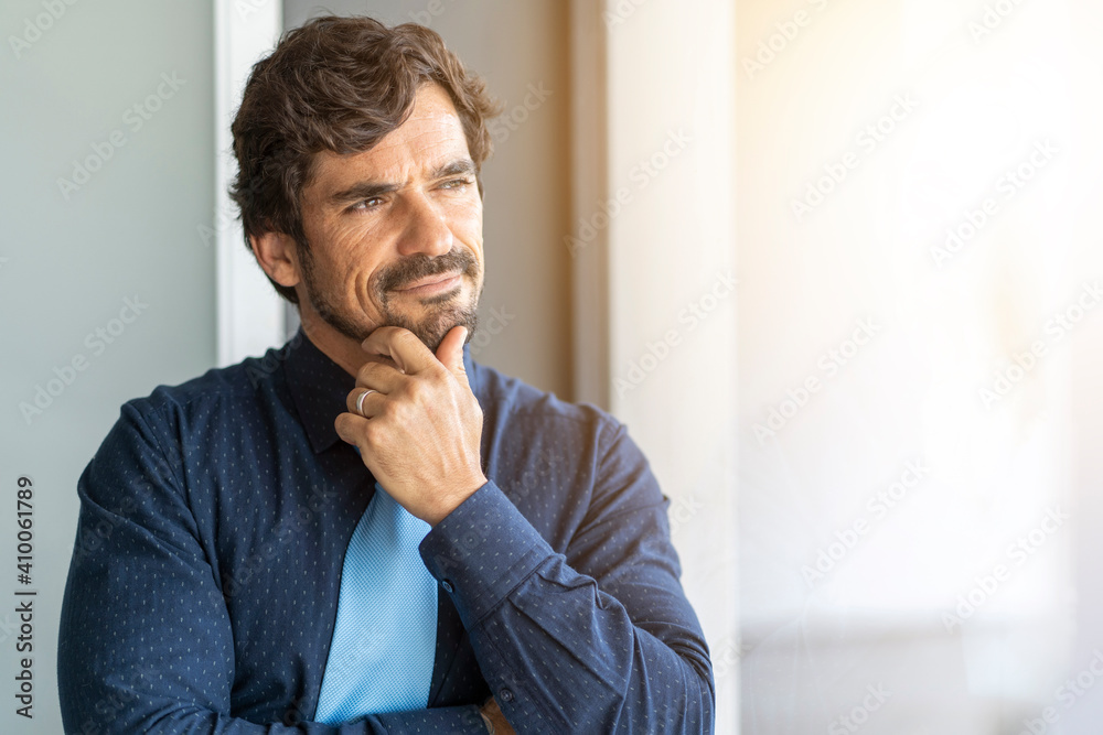 Man standing with hand on his chin looking through the window. Successful male portrait with a happy thinking expression face Man standing with hand on his chin looking through the window. Successful male portrait with a happy thinking expression face
