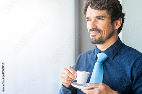Businessman smiling and holding a cup of coffee looking through the window. Successful male portrait with happy face