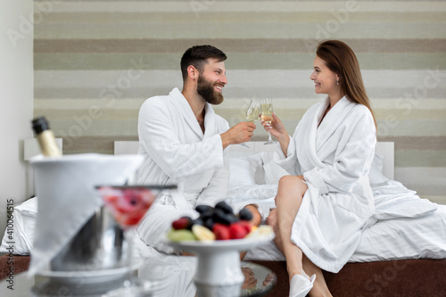 Happy couple a man and a woman in a white coat smile at each other and drink sparkling wine in bed in a hotel room.
