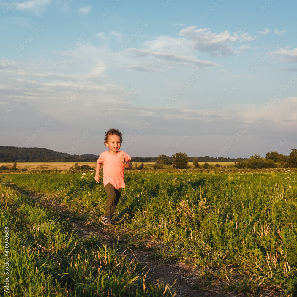 Little curly boy in orange t-shirt running in a meadow, sunset light. Beautiful landscape in summer, very light and happy scene.