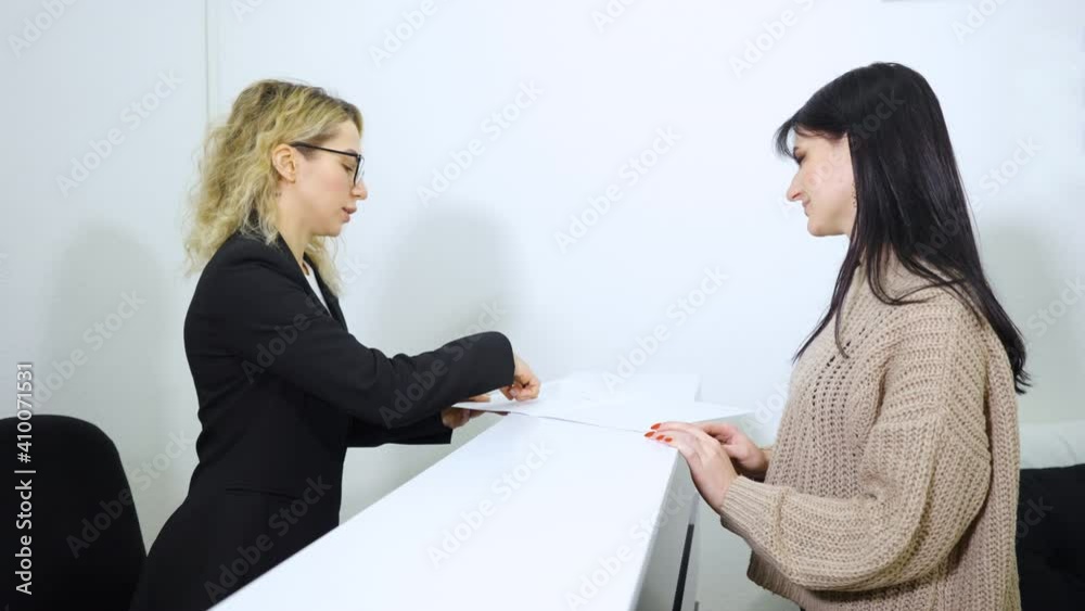 Blonde girl in eyeglasses and black jacket standing at counter and showing instruction list to female visitor. Receptionist informing guest of provided services. Concept of hospitality