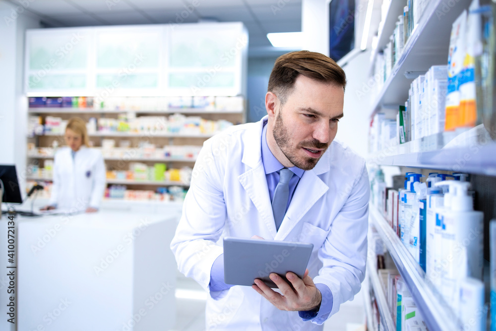 Professional handsome male pharmacist in white coat holding tablet and looking at medicines in pharmacy store or drugstore. Healthcare and apothecary.