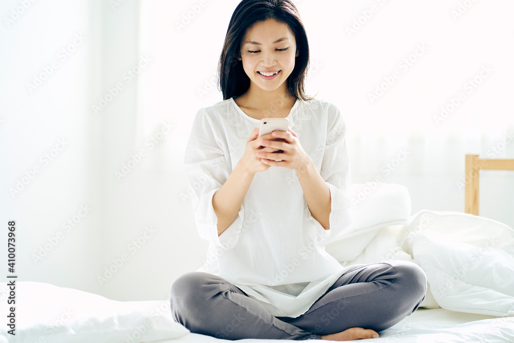 ベッドの上に座りスマートフォンを見る30代日本人女性 ベッドの上に座りスマートフォンを見る30代日本人女性