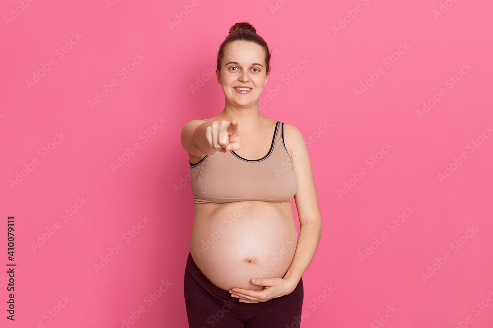 Young pregnant woman pointing finger to camera and touching her bare belly with other hand, looks happy, has hair bun, dresses leggins and tank top, standing isolated over pink background. Young pregnant woman pointing finger to camera and touching her bare belly with other hand, looks happy, has hair bun, dresses leggins and tank top, standing isolated over pink background.
