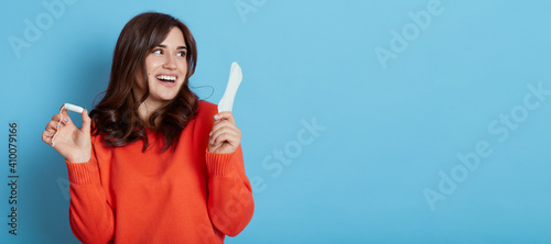 Satisfied lovely woman with dark hair, holds sanitary napkin, uses hygienic product during critical days, smiles positively, looking aside, wearing casual sweater, isolated on blue wall.