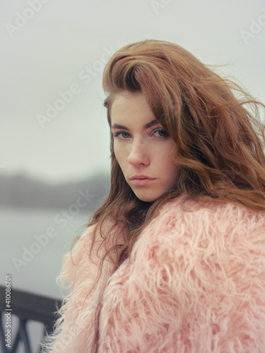 Serious beautiful redhead woman outdoors close up woman portrait in nostalgis film stock color grading posing on river pier in pink fur coat cloudy weather cold season photo