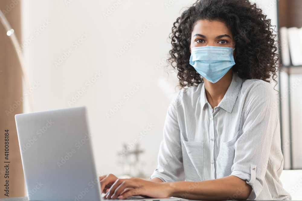 Pretty young African American female freelancer entrepreneur with curly hair wearing a protective medical face mask sitting at the desk in a home office and working on a laptop, looking at the camera Pretty young African American female freelancer entrepreneur with curly hair wearing a protective medical face mask sitting at the desk in a home office and working on a laptop, looking at the camera