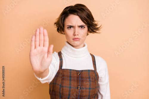 Photo of young serious unhappy frowning girl showing no stop sign in checkered overall isolated on pastel color background