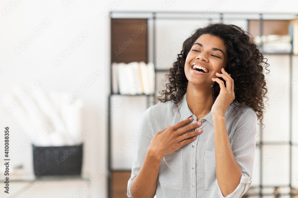 Happy blessed African American teenage girl with curly hair talking on mobile phone and laughing, expressing positive emotions, or a young joyful businesswoman is having a pleasant conversation Happy blessed African American teenage girl with curly hair talking on mobile phone and laughing, expressing positive emotions, or a young joyful businesswoman is having a pleasant conversation