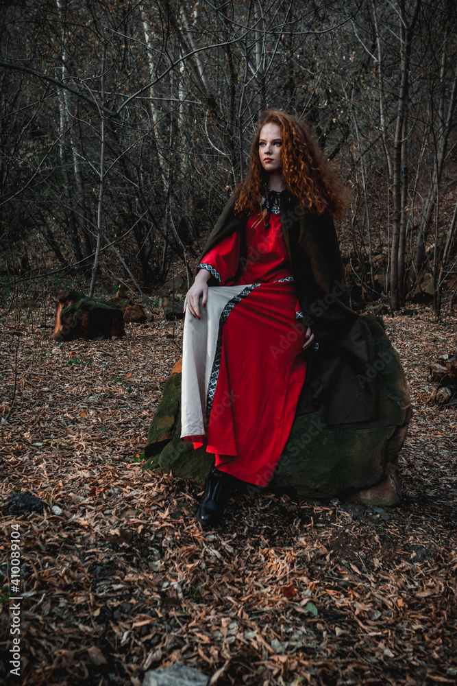 Red-haired woman in a red dress in a historical Celtic costume in the autumn forest Red-haired woman in a red dress in a historical Celtic costume in the autumn forest
