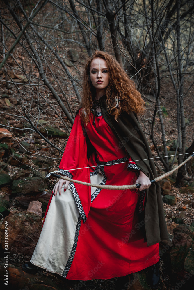 Red-haired woman in a red dress in a historical Celtic costume in the autumn forest Red-haired woman in a red dress in a historical Celtic costume in the autumn forest