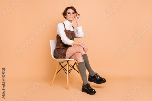 Full size photo of optimistic short hairdo girl sit touch spectacles wear dress socks shoes isolated on peach color background