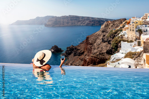 A woman with hat and a drink in her hand enjoys the view from an infinty swimming pool to the beautiful village of Oia on Santorini island, Greece photo