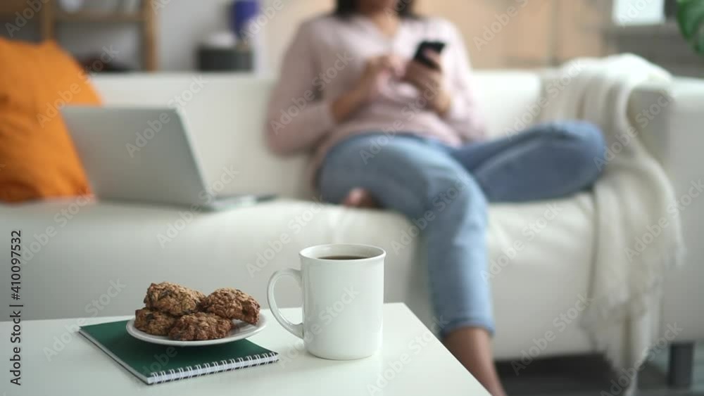 Woman using smartphone and sitting on sofa in home room during pandemic spbd. Young american businesswoman looks at screen of device and holds it her hands, sits on couch in light interior. African Woman using smartphone and sitting on sofa in home room during pandemic spbd. Young american businesswoman looks at screen of device and holds it her hands, sits on couch in light interior. African