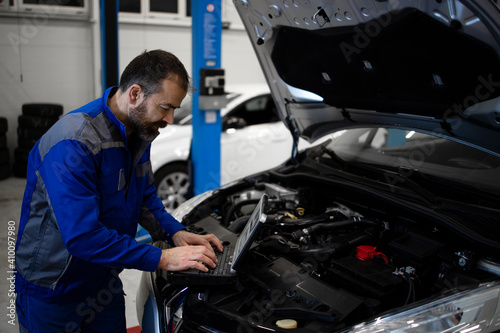 Professional middle aged caucasian car mechanic with laptop computer diagnostic tool standing by vehicle engine area with hood open detecting malfunction.