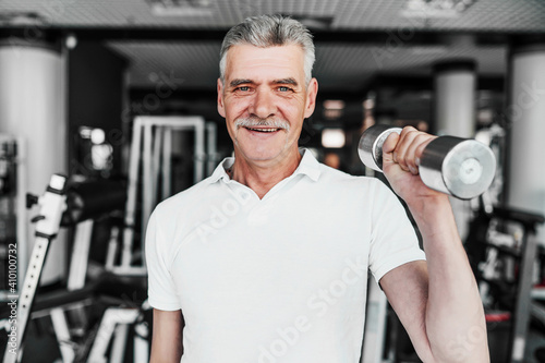 Close up of a smiling senior man in the middle of the gym with a dumbbell in his hands. Training, health