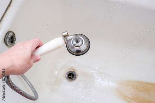 A hand holding shower head with running water against rusty bathtube with limescale, bathroom clining and decalcifying photo