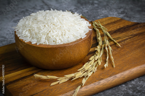 Jasmine rice in a wooden bowl placed on a dark wooden cutting board with rice stalks, rice and jasmine rice in a bowl.
