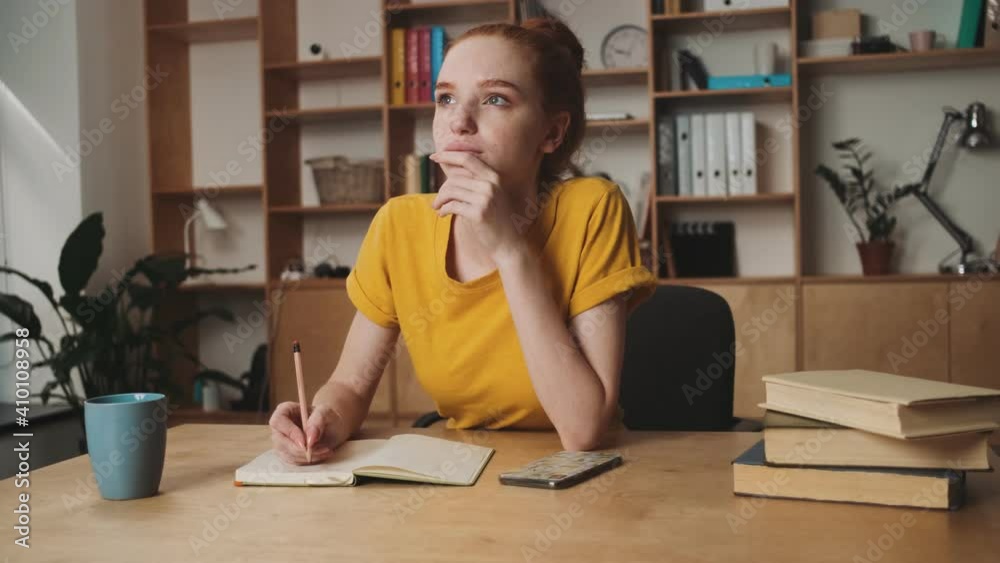 A thinking young redhead woman is writing something sitting at the table in the office