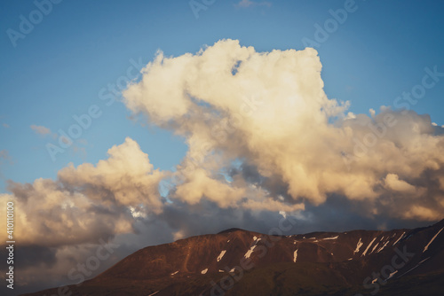 Atmospheric mountain scenery with yellow clouds in blue sky. Scenic landscape with illuminating sunset clouds above mountains with snow. Beautiful sunrise in mountains. Snow on rocks in sunlight.