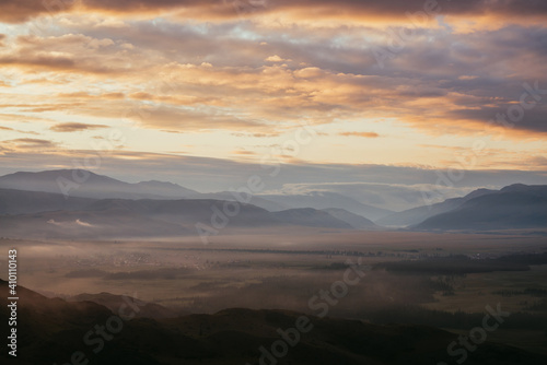 Scenic mountain landscape with golden low clouds above village among mountains silhouettes under dawn cloudy sky. Atmospheric alpine scenery of countryside in low clouds in sundown illuminating color.
