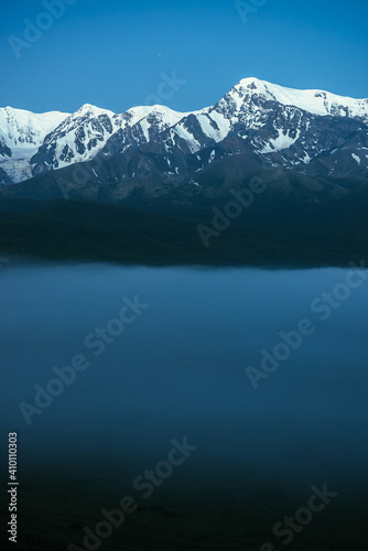 Atmospheric mountains landscape with dense fog and great snow mountain top under twilight sky. Alpine scenery with big snowy mountains over thick fog in night. High snow pinnacle above clouds in dusk.