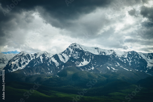 Dramatic mountains landscape with big snowy mountain ridge above sunlit forest in overcast weather. Atmospheric highland scenery with high mountain range under lead gray clouds and sunlight on forest. © Daniil
