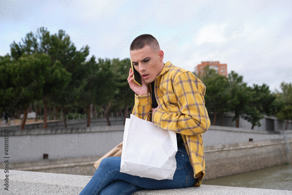 Fototapeta premium Handsome young man wearing make up, concerned speaking on his smartphone, carrying shopping bags. Non binary androgynous guy.