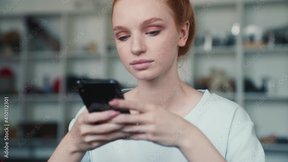 A calm redhead woman is using her smartphone sitting inside the apartments A calm redhead woman is using her smartphone sitting inside the apartments