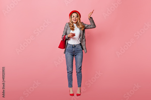 Curly blond woman with glasses and red beret shrugs. Girl in jeans and plaid jacket posing with small bag and telephone in pink studio