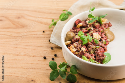 Beef tartare with fresh pea sprout leaves in sauce on a flat plate on a light wooden background.
