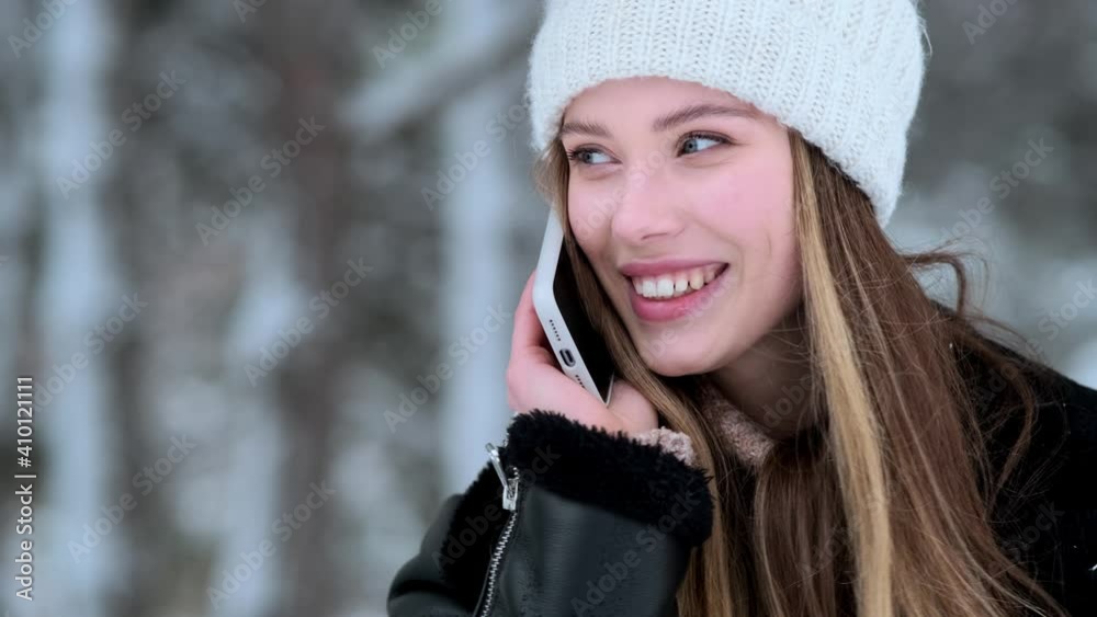 A close-up view of a beautiful young woman wearing winter hat is talking on her smartphone walking outside at wintertime A close-up view of a beautiful young woman wearing winter hat is talking on her smartphone walking outside at wintertime