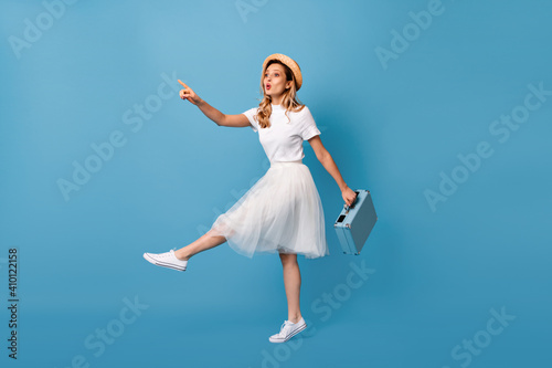 Traveler girl in white fluffy skirt and t-shirt points her finger into distance and holds blue briefbag
