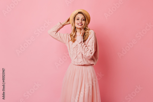 Cute lady in gently pink knitted sweater and fluffy skirt posing on pink background. Woman in boater smiles