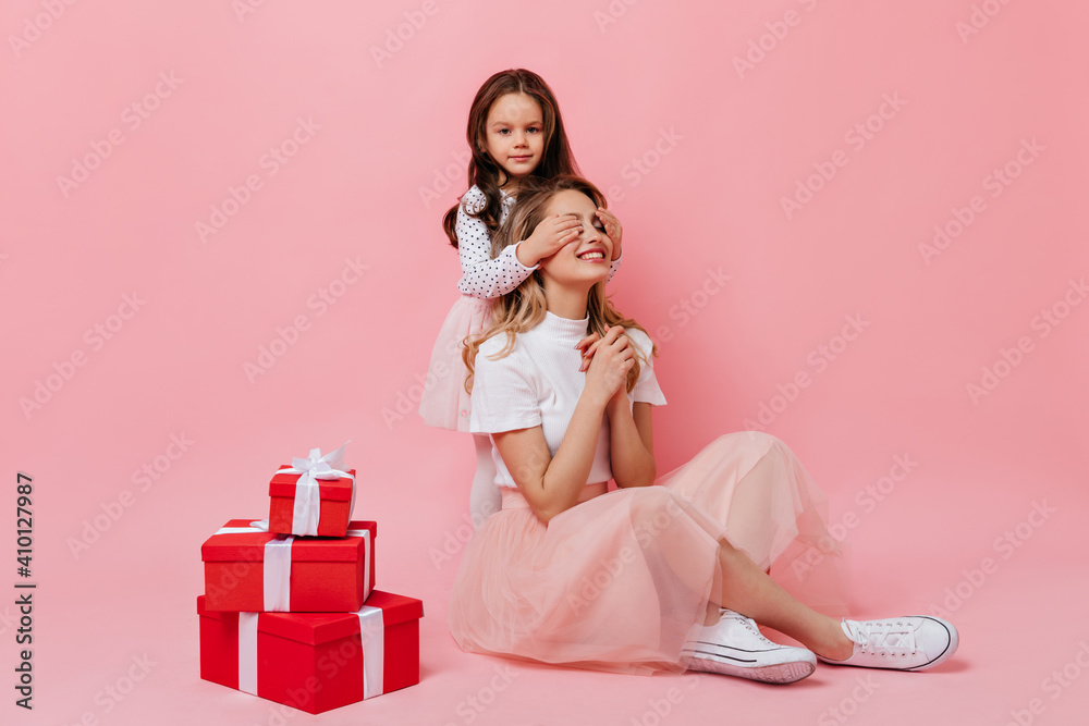 Lady in pink midi skirt and white t-shirt is sitting on pink background while little girl closes her eyes for surprise Lady in pink midi skirt and white t-shirt is sitting on pink background while little girl closes her eyes for surprise